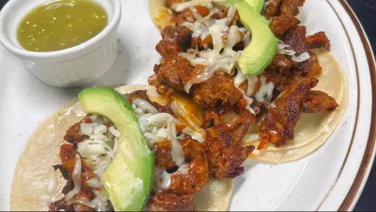 Two open faced tacos al carbon with marinated steak, melted cheese and sliced avocado, with a ramekin of green salsa