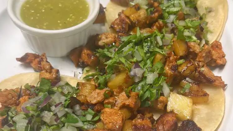 Open faced al pastor tacos with grilled pineapple, cilantro and onion beside a ramekin of green tomatillo salsa
