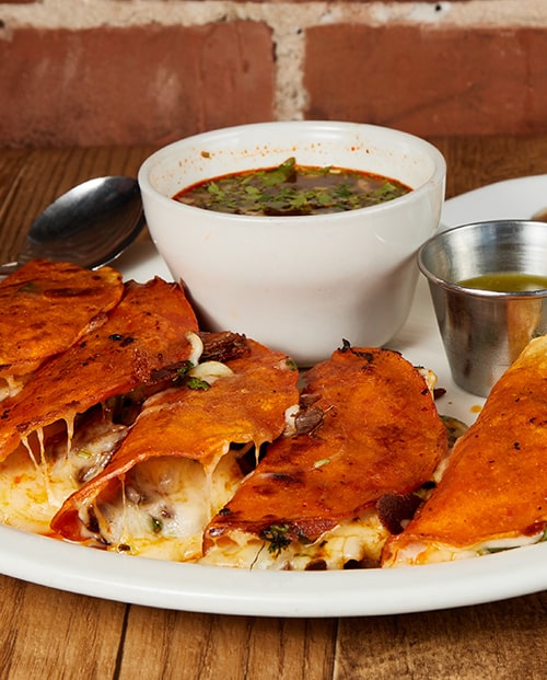 Birria quesa tacos with melting cheese beside a white bowl of consome, on wood against exposed brick