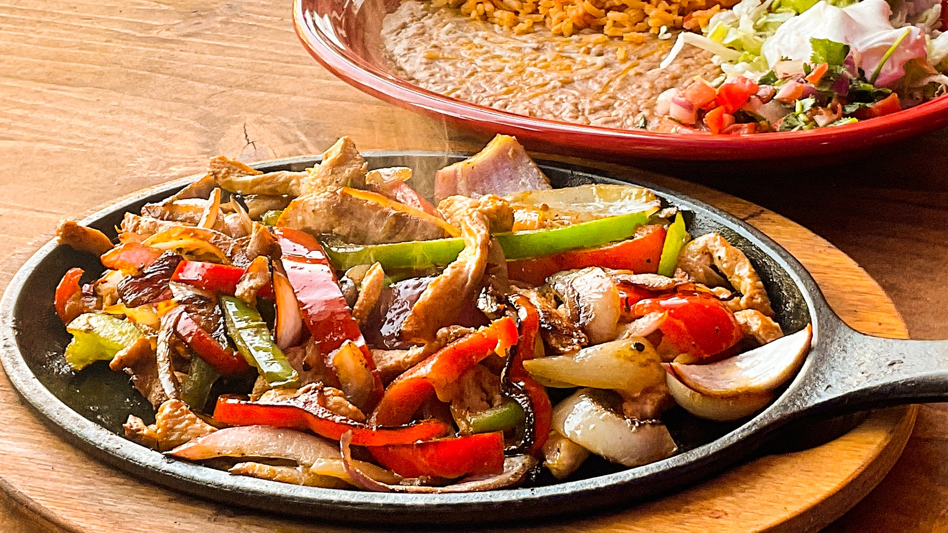 Chicken fajitas with red and green peppers and onions steaming on a black cast iron comal, a red plate of rice, beans, lettuce and pico behind it