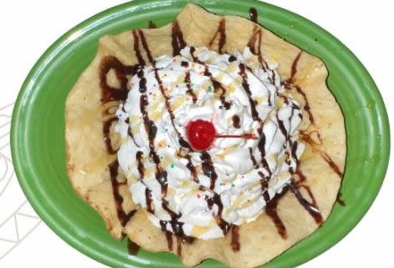 Fried ice cream on a green plate, a dome of whipped cream in a fried tortilla bowl, drizzled with chocolate and topped with a cherry